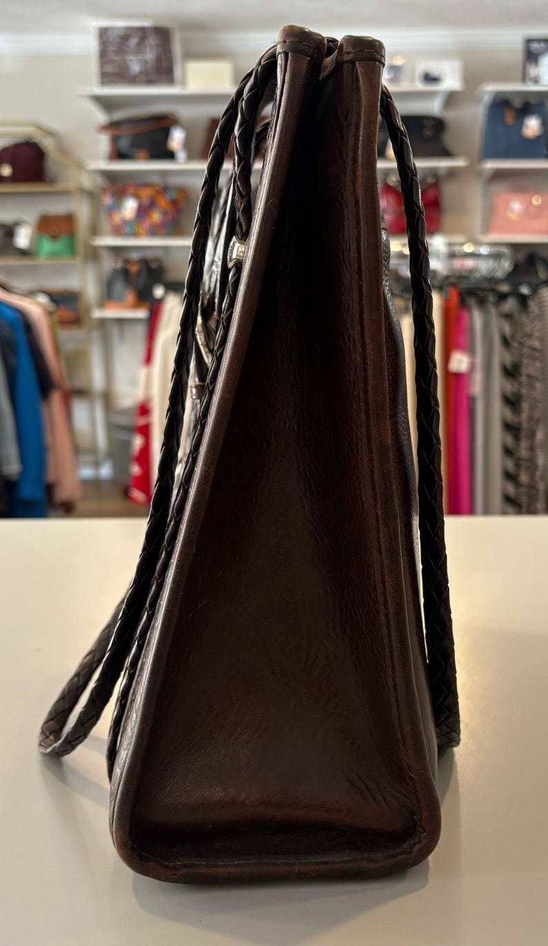May include: A dark brown leather tote bag with braided handles. The bag has a rectangular shape and a textured surface. The handles are a darker shade of brown. The bag is displayed on a white surface.