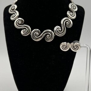 以下が含まれることがあります： シルバーカラーのネックレスとイヤリングのセット。ジュエリーは、繰り返しの渦巻き模様が特徴です。ネックレスは黒いマネキンに、イヤリングは透明なスタンドに展示されています。背景はニュートラルグレーです。