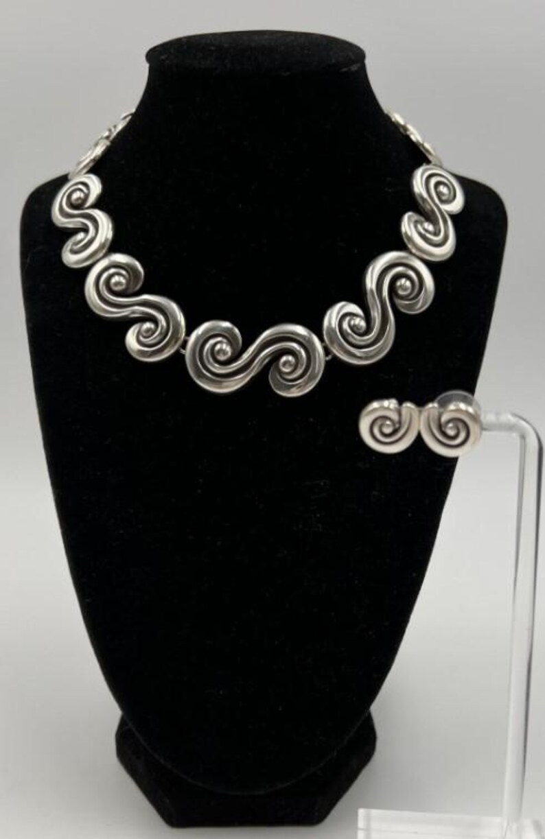 以下が含まれることがあります： シルバーカラーのネックレスとイヤリングのセット。ジュエリーは、繰り返しの渦巻き模様が特徴です。ネックレスは黒いマネキンに、イヤリングは透明なスタンドに展示されています。背景はニュートラルグレーです。