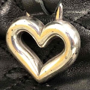 Pu&ograve; includere: Un ciondolo a forma di cuore in argento con una finitura liscia e lucida. Il ciondolo &egrave; attaccato a una borsa in pelle nera.