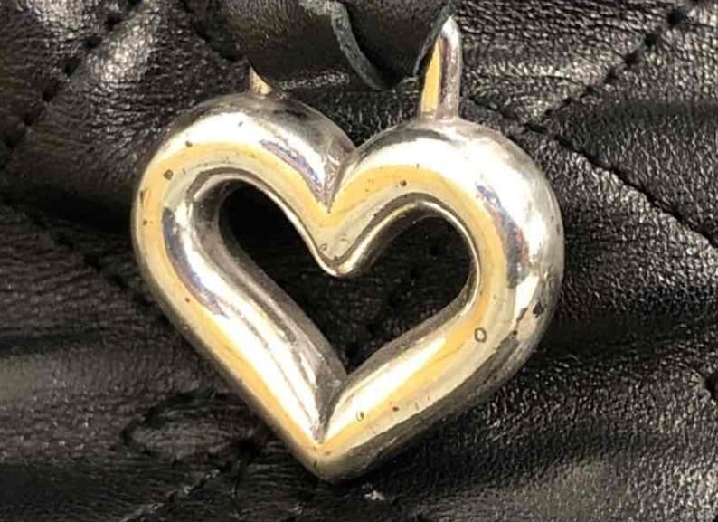 Pu&ograve; includere: Un ciondolo a forma di cuore in argento con una finitura liscia e lucida. Il ciondolo &egrave; attaccato a una borsa in pelle nera.