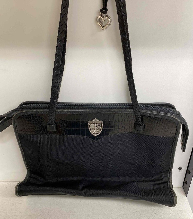 K&ouml;nnte beinhalten: Eine schwarze Ledertasche mit geflochtenem Trageriemen und einem silbernen Herz-Anh&auml;nger. Die Tasche hat ein Kroko-Pr&auml;gemuster und ein silbernes, schildf&ouml;rmiges Logo.