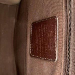 May include: Close-up of a brown leather tag with the Brighton logo and text. The tag is rectangular with rounded corners and is stitched to a brown fabric. The background is a textured brown fabric.