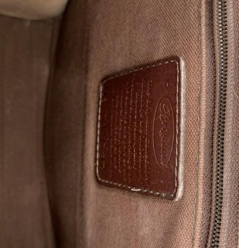 May include: Close-up of a brown leather tag with the Brighton logo and text. The tag is rectangular with rounded corners and is stitched to a brown fabric. The background is a textured brown fabric.