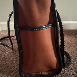 May include: A brown leather tote bag with black trim and white stitching. The bag has a braided handle and a pocket on the front. The bag is sitting on a brown carpet.