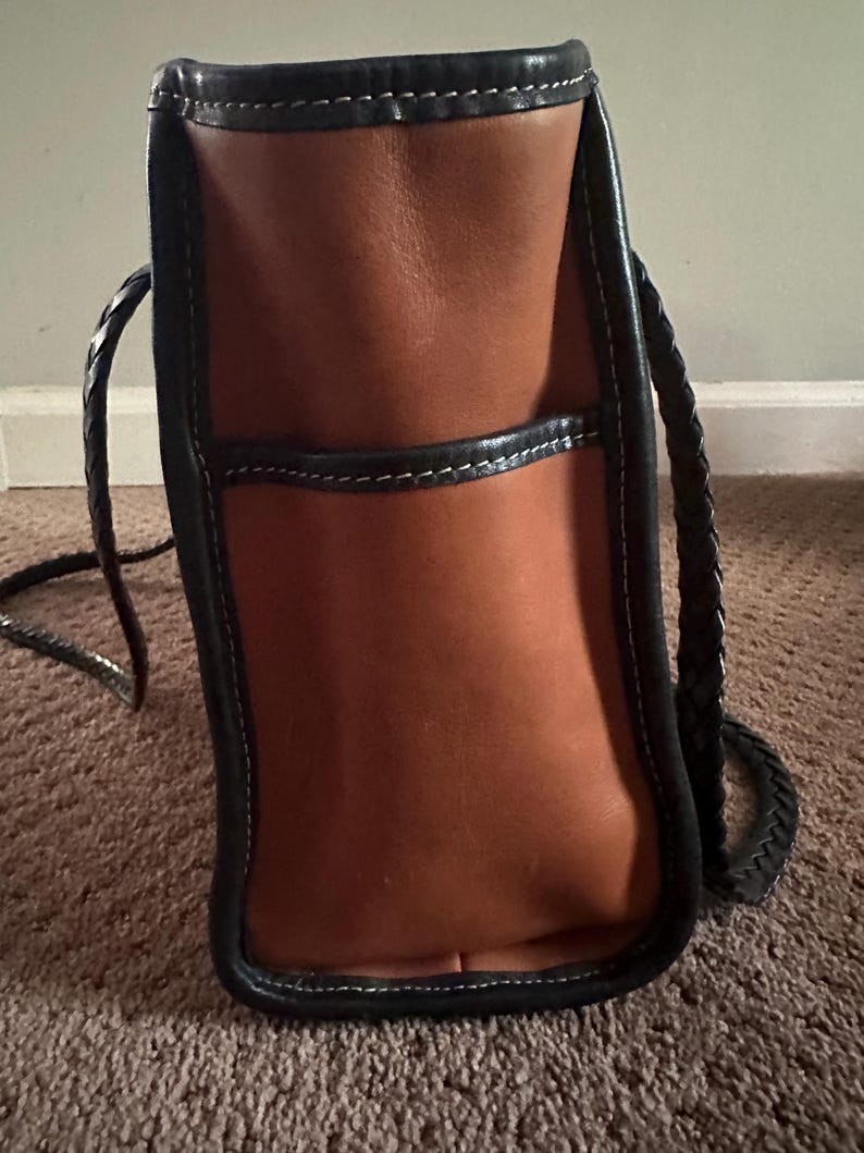 May include: A brown leather tote bag with black trim and white stitching. The bag has a braided handle and a pocket on the front. The bag is sitting on a brown carpet.