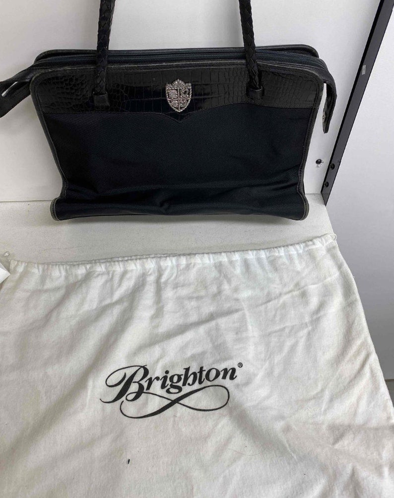 K&ouml;nnte beinhalten: Schwarze Ledertasche mit silbernem Brighton-Logo und einem Staubbeutel mit der Aufschrift "Brighton" in Kursivschrift.