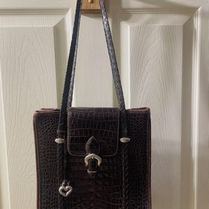 May include: A brown, faux-crocodile leather tote bag with a rectangular shape. The bag features a flap closure with a silver buckle, a braided shoulder strap, and a silver heart charm. The bag is hanging on a wooden hook.