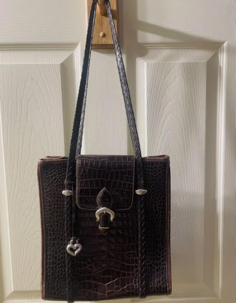 May include: A brown, faux-crocodile leather tote bag with a rectangular shape. The bag features a flap closure with a silver buckle, a braided shoulder strap, and a silver heart charm. The bag is hanging on a wooden hook.