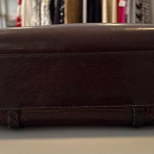 May include: A dark brown leather handbag with a rectangular shape. The bag has a textured surface and a stitched design along the top and bottom. The bottom features a crocodile-like pattern and braided accents.
