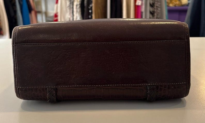 May include: A dark brown leather handbag with a rectangular shape. The bag has a textured surface and a stitched design along the top and bottom. The bottom features a crocodile-like pattern and braided accents.