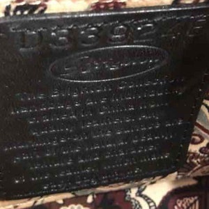 Pu&ograve; includere: Primo piano di un'etichetta in pelle nera con il testo "Dooney & Bourke" e un testo pi&ugrave; piccolo "The Signature Collection" in bianco. L'etichetta &egrave; attaccata a un tessuto con un motivo paisley.