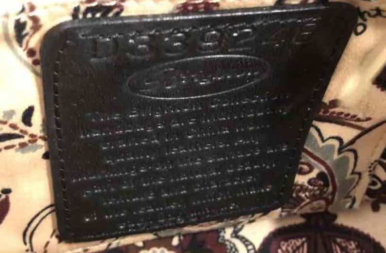 Pu&ograve; includere: Primo piano di un'etichetta in pelle nera con il testo "Dooney & Bourke" e un testo pi&ugrave; piccolo "The Signature Collection" in bianco. L'etichetta &egrave; attaccata a un tessuto con un motivo paisley.