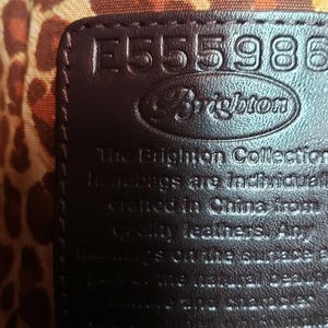 May include: Close-up of a dark brown leather tag with the number "E555986" embossed on it. The Brighton logo is visible. The tag is set against a leopard print fabric background. Text on the tag reads "The Brighton Collection handbags are individually crafted in China from quality leathers."
