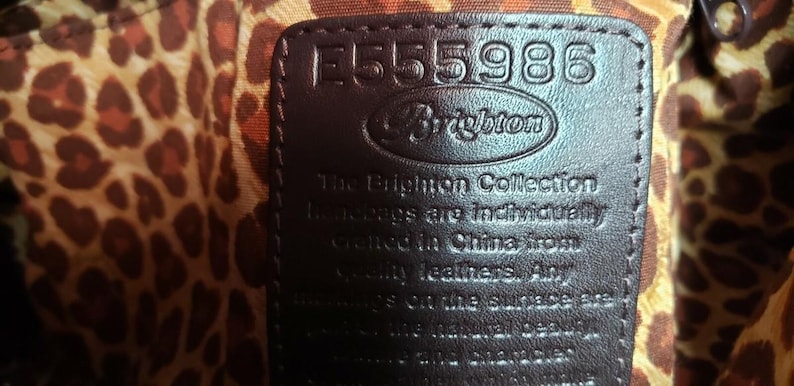 May include: Close-up of a dark brown leather tag with the number "E555986" embossed on it. The Brighton logo is visible. The tag is set against a leopard print fabric background. Text on the tag reads "The Brighton Collection handbags are individually crafted in China from quality leathers."