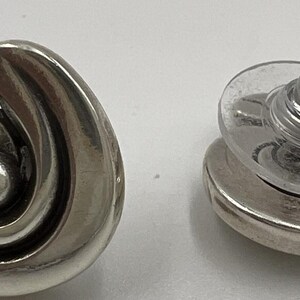 以下が含まれることがあります： シルバーカラーのスタッドピアス。片方のイヤリングはスパイラルデザインで、もう片方はポストと裏側を示しています。イヤリングは研磨仕上げで、シンプルでエレガントなスタイルです。