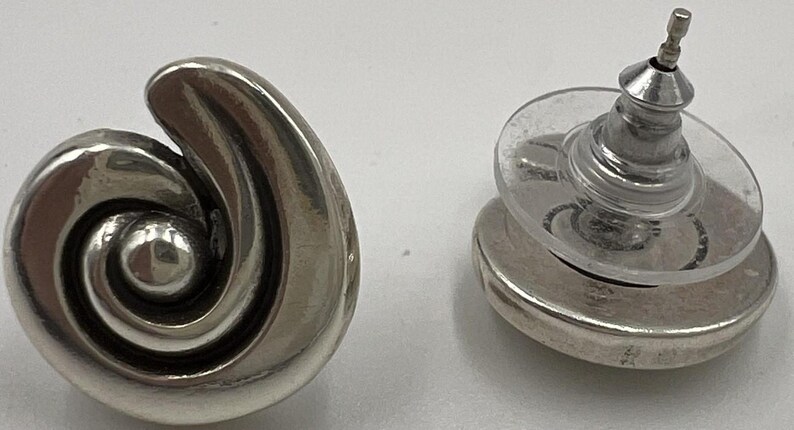 以下が含まれることがあります： シルバーカラーのスタッドピアス。片方のイヤリングはスパイラルデザインで、もう片方はポストと裏側を示しています。イヤリングは研磨仕上げで、シンプルでエレガントなスタイルです。