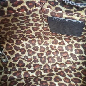 May include: Interior view of a brown handbag with a leopard print lining. The bag has a dark brown leather handle and a small leather tag. A silver zipper and clasp are visible.