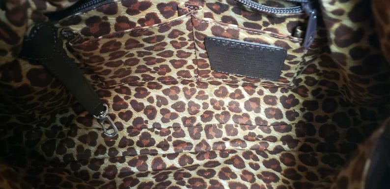 May include: Interior view of a brown handbag with a leopard print lining. The bag has a dark brown leather handle and a small leather tag. A silver zipper and clasp are visible.