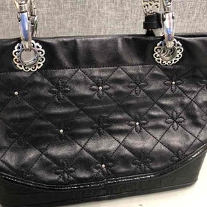 Pu&ograve; includere: Borsa tote in pelle nera con dettagli in argento e un design trapuntato con piccoli accenti floreali.