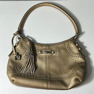 May include: Gold-colored leather shoulder bag with a curved shape and a single handle. The bag features a decorative tassel, a silver-tone metal accent, and stitched detailing. The leather has a textured appearance.