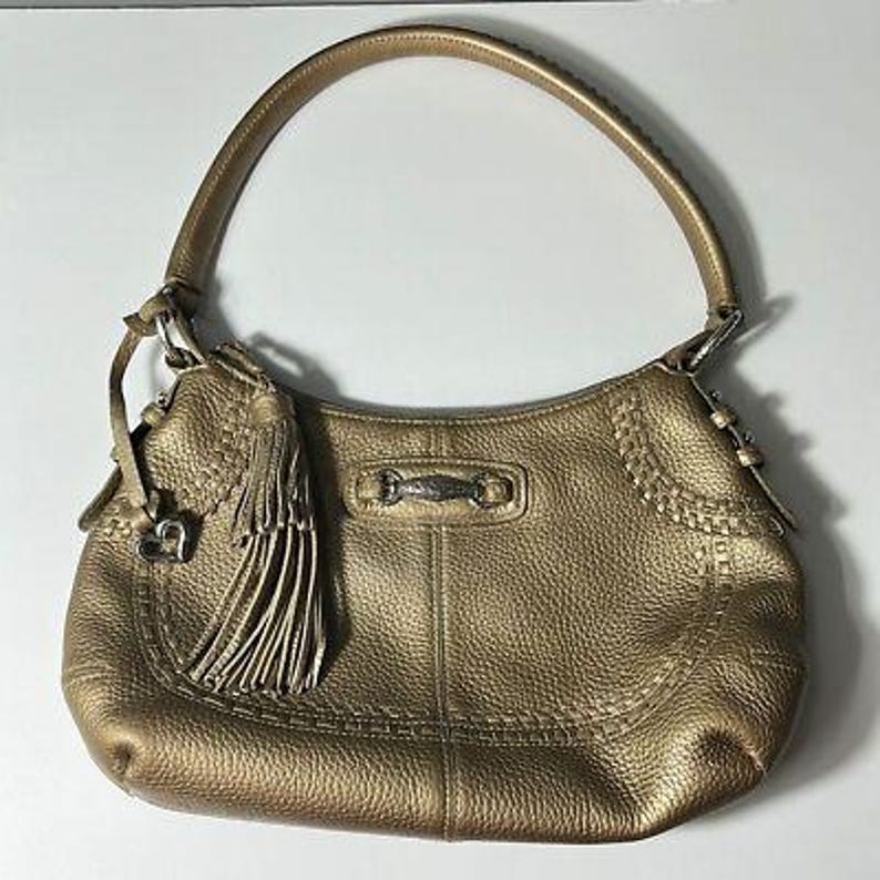 May include: Gold-colored leather shoulder bag with a curved shape and a single handle. The bag features a decorative tassel, a silver-tone metal accent, and stitched detailing. The leather has a textured appearance.