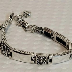 Puede incluir: Pulsera plateada con un dise&ntilde;o ornamentado. Presenta eslabones rectangulares con patrones grabados y un cierre decorativo. Peque&ntilde;os dijes de campana cuelgan del extremo, a&ntilde;adiendo un toque delicado. La pulsera est&aacute; hecha de un metal brillante y reflectante.