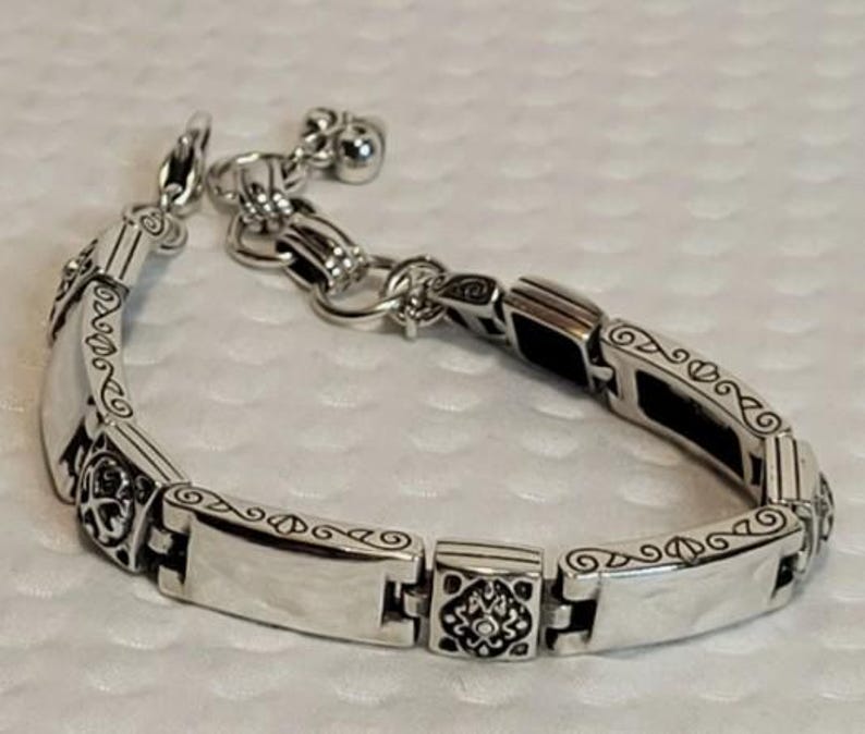 Puede incluir: Pulsera plateada con un dise&ntilde;o ornamentado. Presenta eslabones rectangulares con patrones grabados y un cierre decorativo. Peque&ntilde;os dijes de campana cuelgan del extremo, a&ntilde;adiendo un toque delicado. La pulsera est&aacute; hecha de un metal brillante y reflectante.