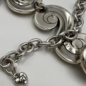 以下が含まれることがあります： 渦巻き模様の装飾的なリンクとハート型のチャームが付いたシルバーカラーのブレスレット。チャームには「B」と「J」のイニシャルが刻まれています。ブレスレットは一連の楕円形のリンクで接続されています。