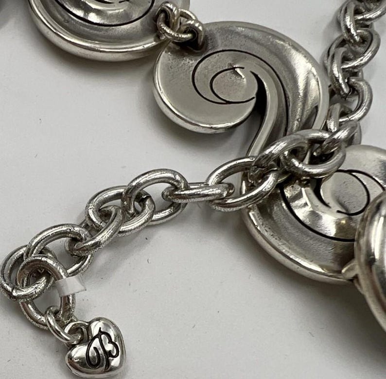 以下が含まれることがあります： 渦巻き模様の装飾的なリンクとハート型のチャームが付いたシルバーカラーのブレスレット。チャームには「B」と「J」のイニシャルが刻まれています。ブレスレットは一連の楕円形のリンクで接続されています。