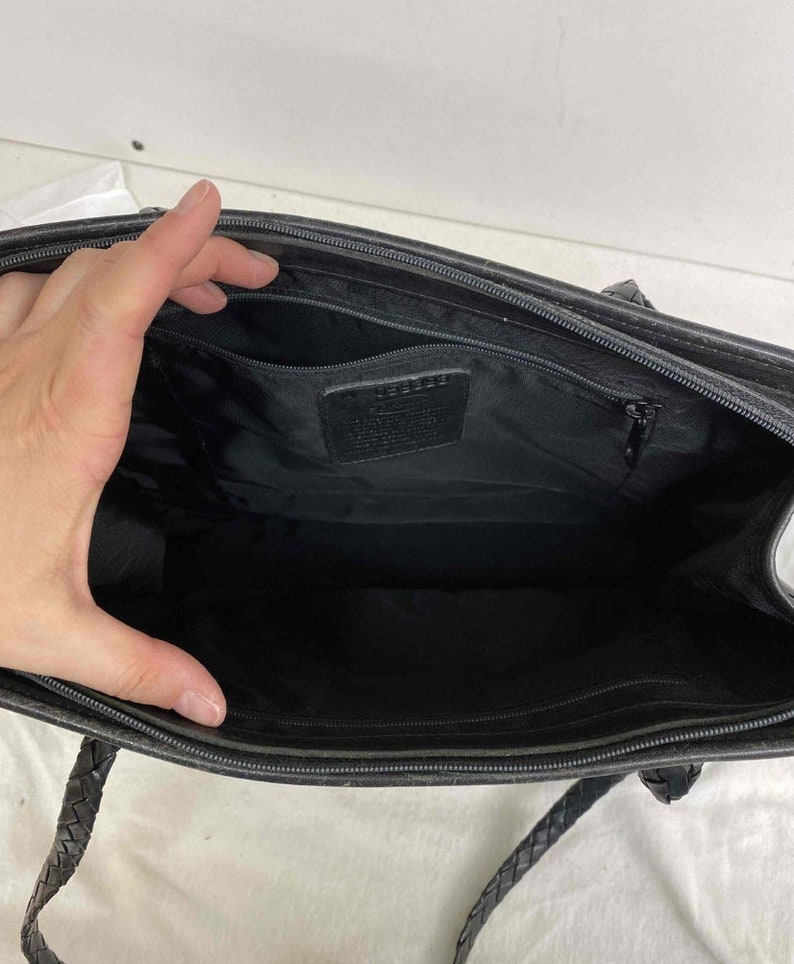 K&ouml;nnte beinhalten: Schwarze Ledertasche mit geflochtenem Trageriemen und einer Rei&szlig;verschlusstasche. Die Tasche hat ein schwarzes Stofffutter und ein Coach-Etikett innen.