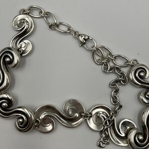 以下が含まれることがあります： 銀色のネックレス。一連の様式化された渦巻きスクロールデザインが特徴です。ネックレスは、調節可能なチェーンとロブスタークラスプ留めを備えています。デザインは対称的で、スクロール要素がネックレスの長さに沿って均等に配置されています。