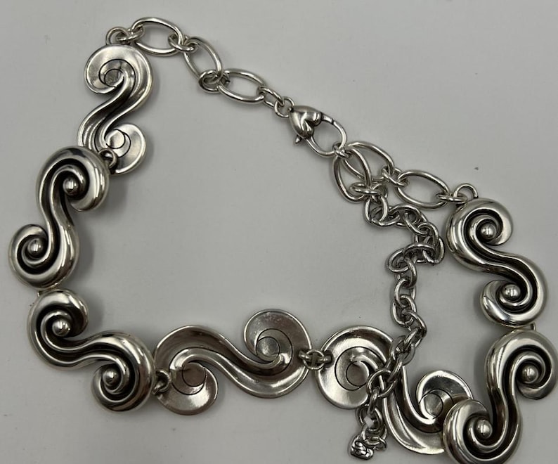 以下が含まれることがあります： 銀色のネックレス。一連の様式化された渦巻きスクロールデザインが特徴です。ネックレスは、調節可能なチェーンとロブスタークラスプ留めを備えています。デザインは対称的で、スクロール要素がネックレスの長さに沿って均等に配置されています。