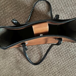 May include: A brown leather tote bag with black braided handles and trim. The bag has a structured design with a central compartment and a small, light brown flap closure. The interior lining features a striped pattern.