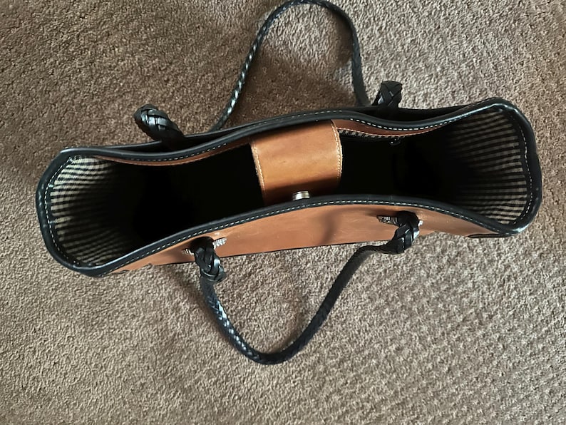 May include: A brown leather tote bag with black braided handles and trim. The bag has a structured design with a central compartment and a small, light brown flap closure. The interior lining features a striped pattern.