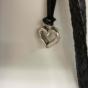 K&ouml;nnte beinhalten: Ein silberner herzf&ouml;rmiger Anh&auml;nger h&auml;ngt an einem schwarzen geflochtenen Lederband.