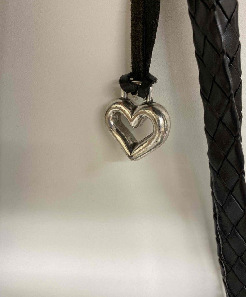 K&ouml;nnte beinhalten: Ein silberner herzf&ouml;rmiger Anh&auml;nger h&auml;ngt an einem schwarzen geflochtenen Lederband.