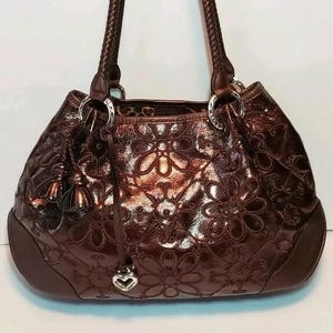 May include: A brown leather handbag with a floral embossed pattern. The bag has braided handles, silver-tone hardware, and a heart-shaped charm. The interior lining is a leopard print. A tassel hangs from the side.