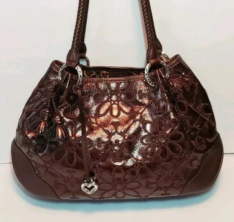 May include: A brown leather handbag with a floral embossed pattern. The bag has braided handles, silver-tone hardware, and a heart-shaped charm. The interior lining is a leopard print. A tassel hangs from the side.