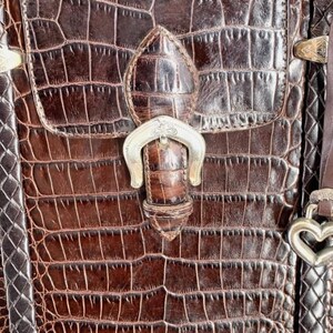 May include: Close-up of a brown leather handbag with a crocodile pattern. The bag features a flap closure with a silver-colored buckle and a decorative heart-shaped clasp. The leather has a textured appearance.
