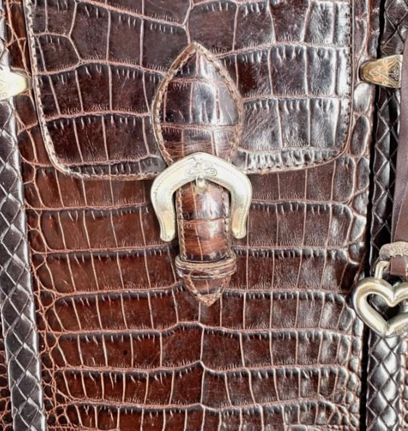 May include: Close-up of a brown leather handbag with a crocodile pattern. The bag features a flap closure with a silver-colored buckle and a decorative heart-shaped clasp. The leather has a textured appearance.