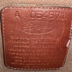 May include: A close-up of a brown leather tag with the brand name "Brighton" embossed in the center. The tag also features the number "054571" and the letter "A". The tag is stitched onto a tan fabric.