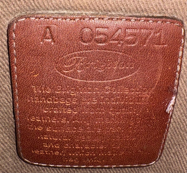 May include: A close-up of a brown leather tag with the brand name "Brighton" embossed in the center. The tag also features the number "054571" and the letter "A". The tag is stitched onto a tan fabric.