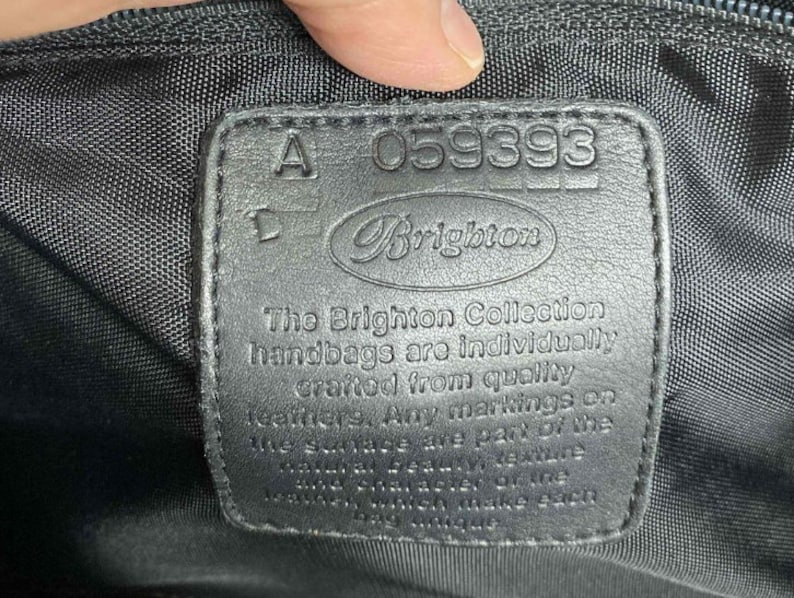 K&ouml;nnte beinhalten: Ein schwarzes Leder-Etikett mit dem Text "A-059393 Brighton" und dem Text "The Brighton Collection handbags are individually crafted from quality leathers. Any markings on the surface are part of the natural beauty of the leather and will make each bag unique."