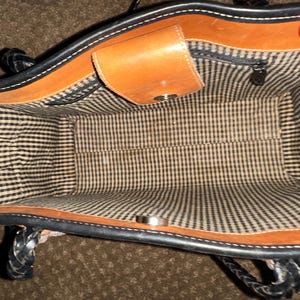 May include: Open view of a brown and black leather handbag. The interior features a black and white checkered lining. The bag has a small, tan leather flap with a silver snap closure. The handles are black and braided.