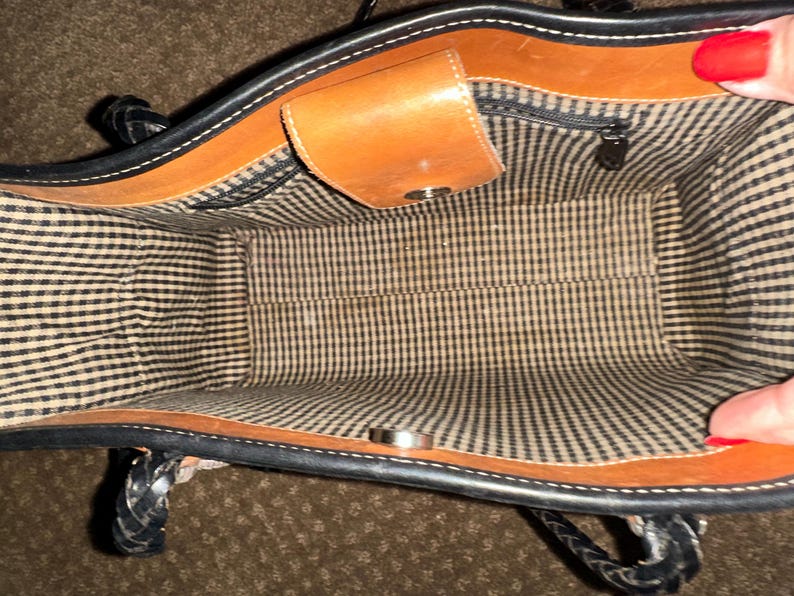 May include: Open view of a brown and black leather handbag. The interior features a black and white checkered lining. The bag has a small, tan leather flap with a silver snap closure. The handles are black and braided.