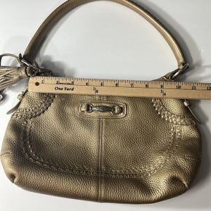 May include: A metallic gold handbag with a top handle and a decorative tassel. The bag has a textured surface and decorative stitching along the edges. The bag measures approximately 12 inches in length.
