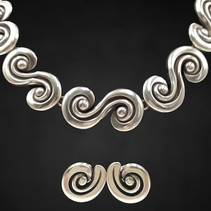 以下が含まれることがあります： シルバーカラーのネックレスとイヤリングのセット。ジュエリーは、繰り返しの渦巻き模様が特徴です。ネックレスは、接続された、様式化された波のような形で構成されています。イヤリングは、より小さな単一の渦巻きでデザインを反映しています。