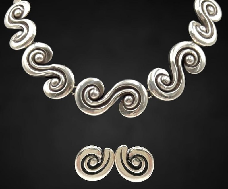 以下が含まれることがあります： シルバーカラーのネックレスとイヤリングのセット。ジュエリーは、繰り返しの渦巻き模様が特徴です。ネックレスは、接続された、様式化された波のような形で構成されています。イヤリングは、より小さな単一の渦巻きでデザインを反映しています。