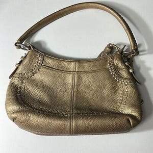 May include: A metallic gold-colored handbag with a curved shape and a single handle. The bag features decorative stitching along the sides and a small tassel. The leather has a textured appearance, and the hardware is silver-toned.